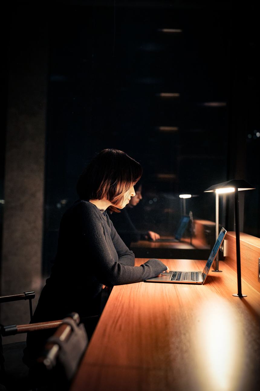 A woman working late on her laptop at a long lamp-lit desk in a dark office