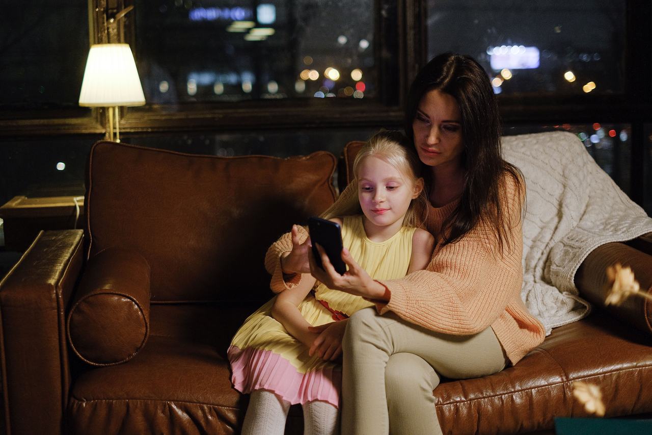 A mother and her young daughter on a sofa in the evening, looking at a phone together by lamp light